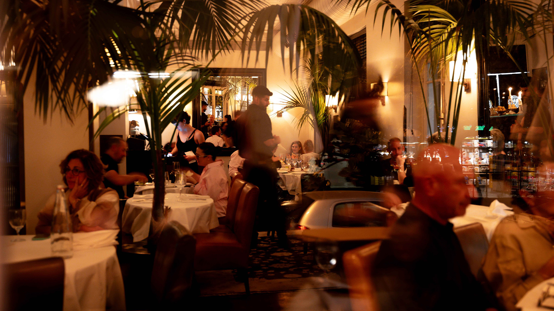 Guests enjoying a lively evening at the Hotel Montefiore restaurant, surrounded by elegant decor, lush palm plants, and warm ambient lighting.